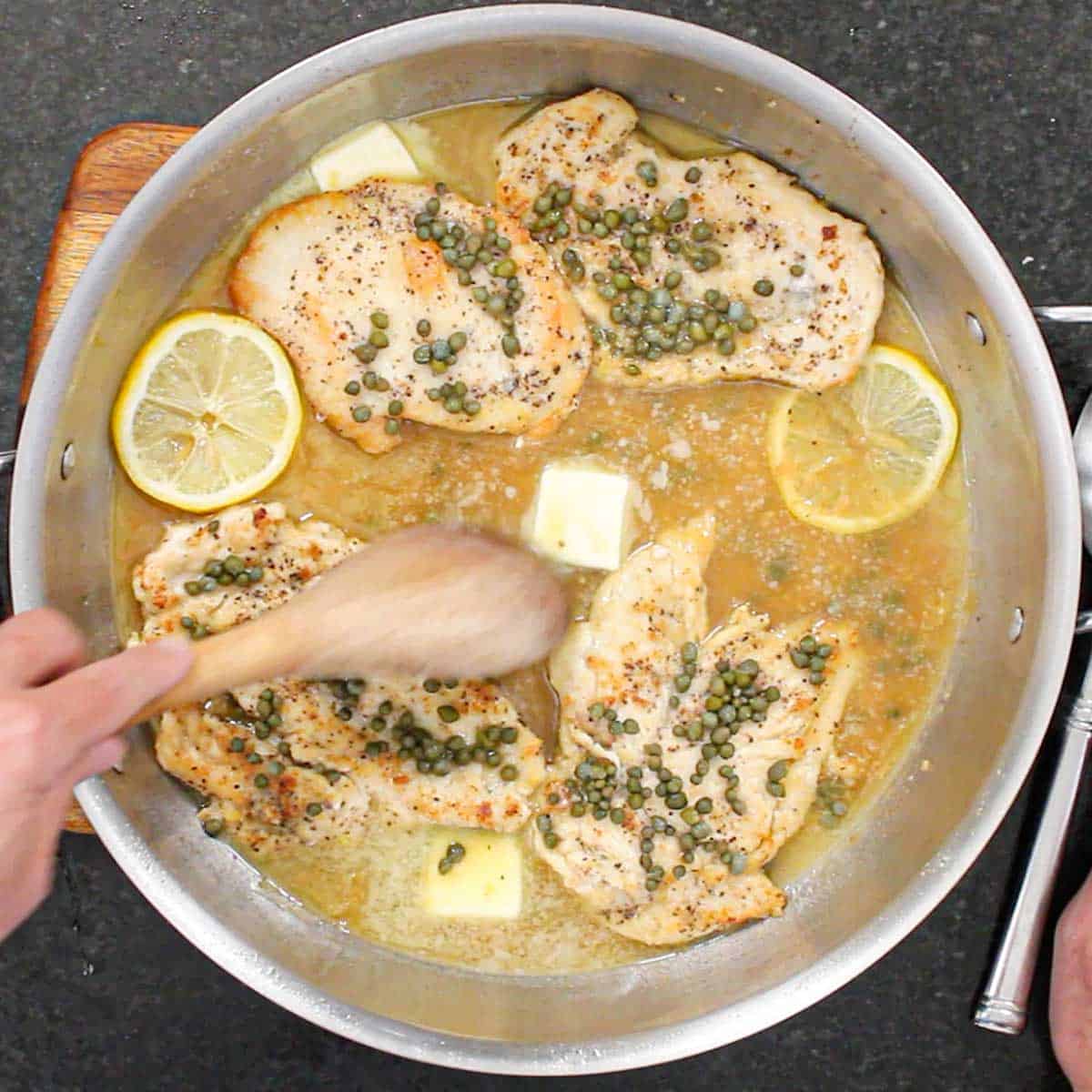 A person using a wooden spoon to stir pads of butter that is in the process of being melted in a pan filled with homemade chicken piccata and lemon wedges.