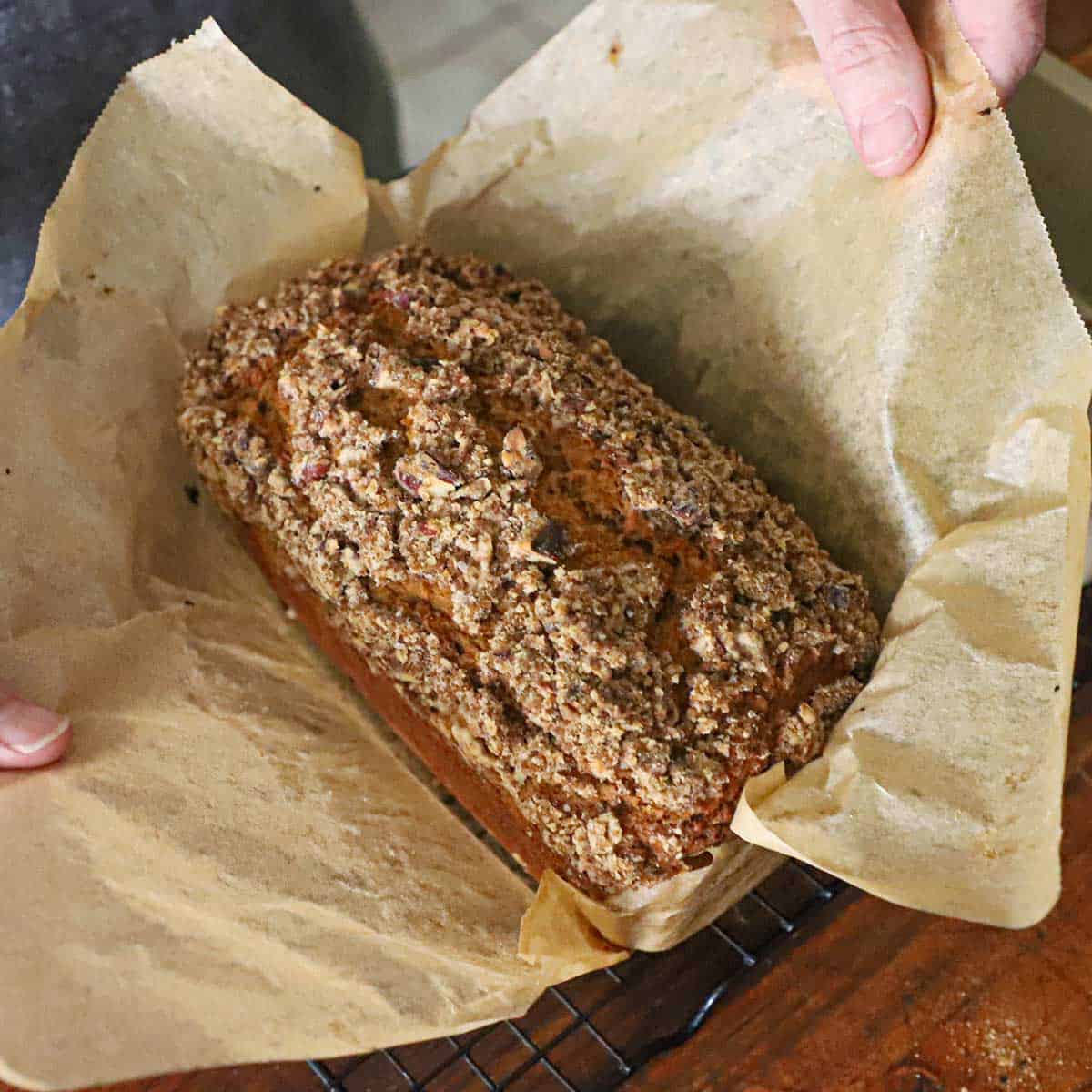 A person pulling brown parchment paper away from a freshly baked best homemade pumpkin bread loaf that is resting on a wire rack.