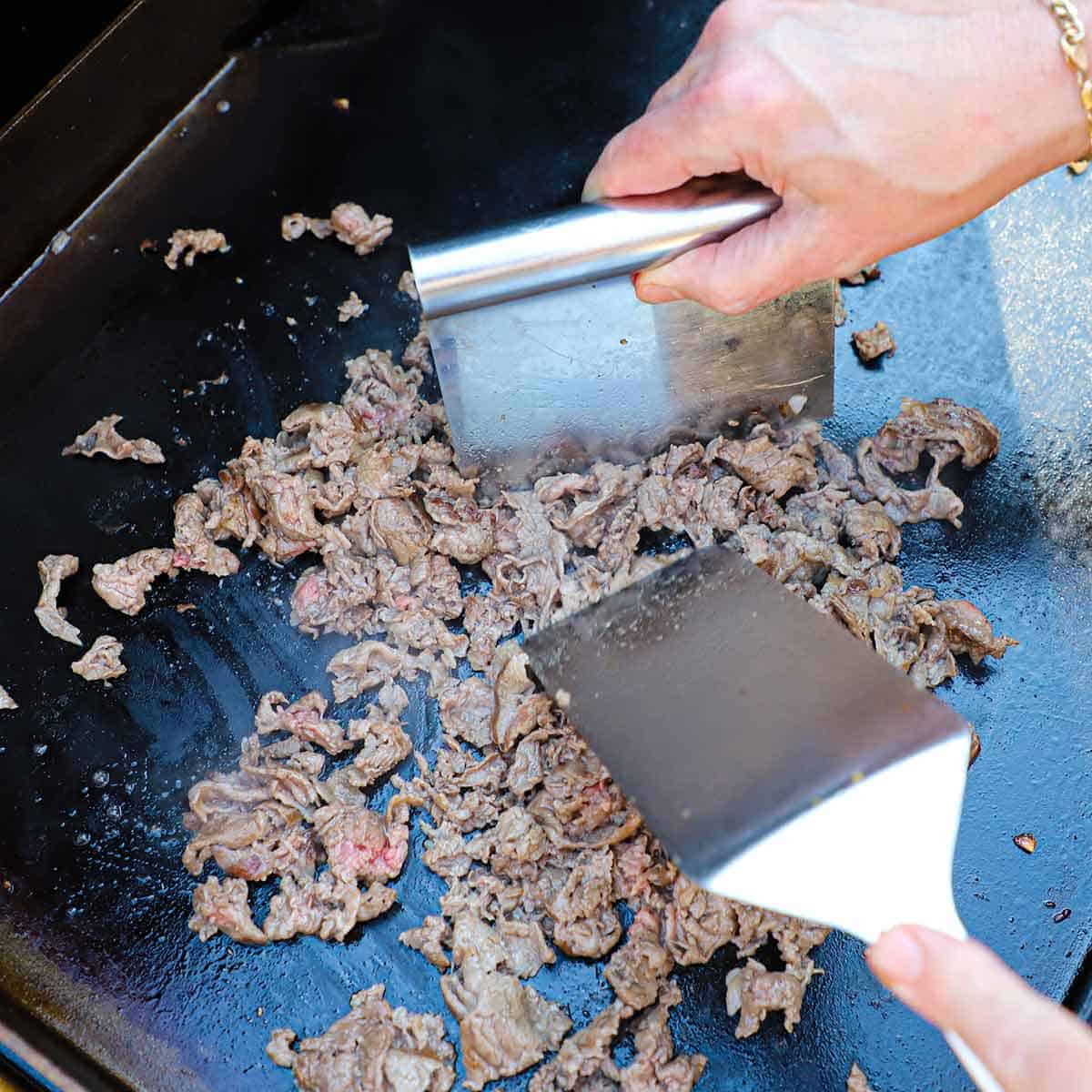 A person using a large silver spatula and a bench scraper to gently shred thinly sliced ribeye that is being cooked on an outdoor griddle.