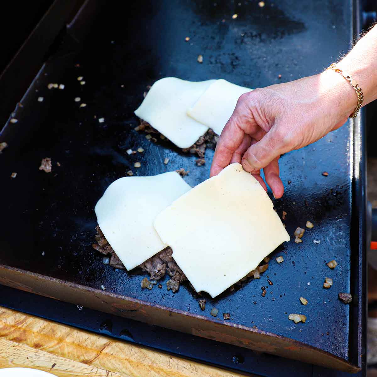 A person placing squares of white American cheese over two mounds of chopped ribeye and onions on a large outdoor griddle.