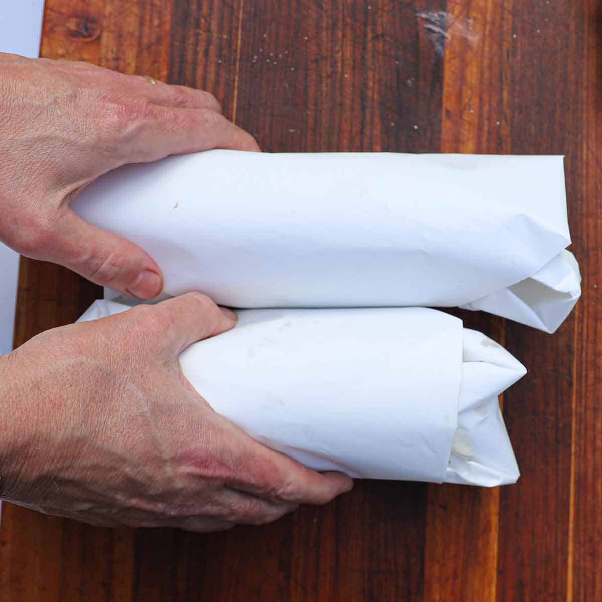 A person placing two homemade Philly cheesesteaks that are wrapped in white butcher paper on a wooden cutting board.