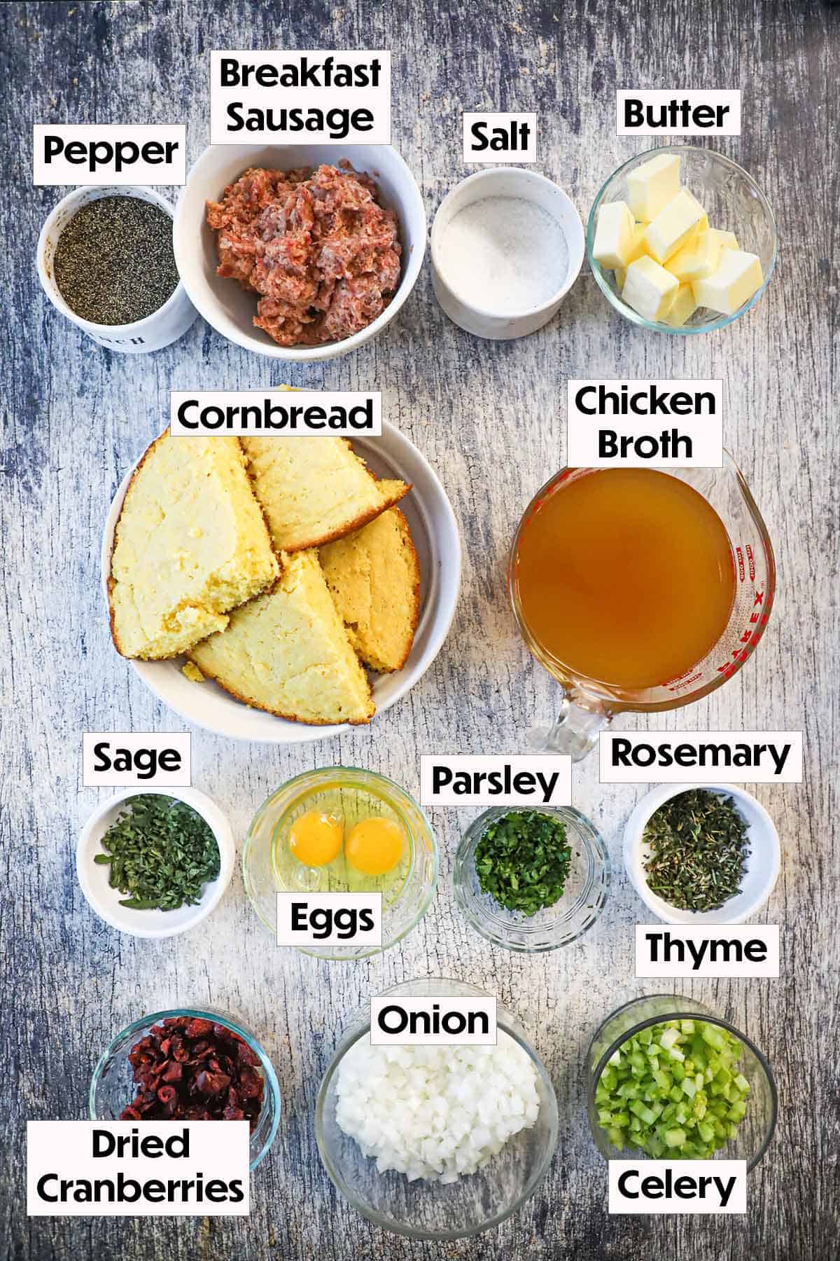 An arrangement of ingredients for cornbread dressing with sausage and cranberries on a grey wooden background including cornbread, chicken broth, sausage, butter, eggs, onion, cranberries, celery, and seasonings.