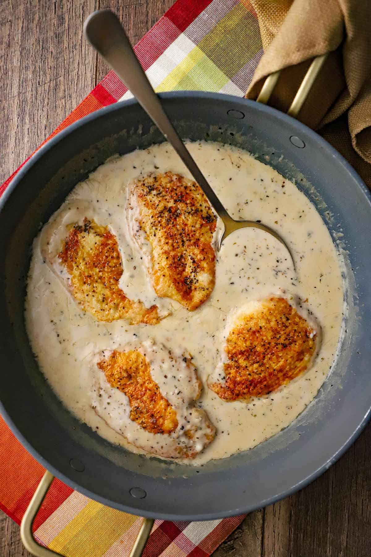 A large grey non-stick skillet filled with four seared chicken breasts in a garlic Parmesan sauce with a large spoon resting in the skillet.