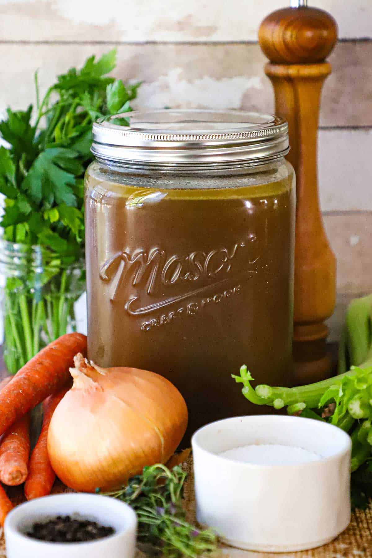 An extra-large Mason jar filled with homemade roasted turkey stock with a glass of Italian parsley, carrots, an onion, a bowl of peppercorns, and a bowl of salt surrounding it.