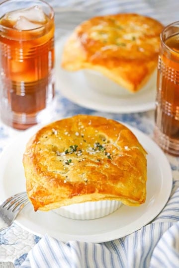 A white plate holding a large ramekin of leftover turkey pot pie with butter puff pastry sitting next to another plate of the same and two glasses of iced tea.