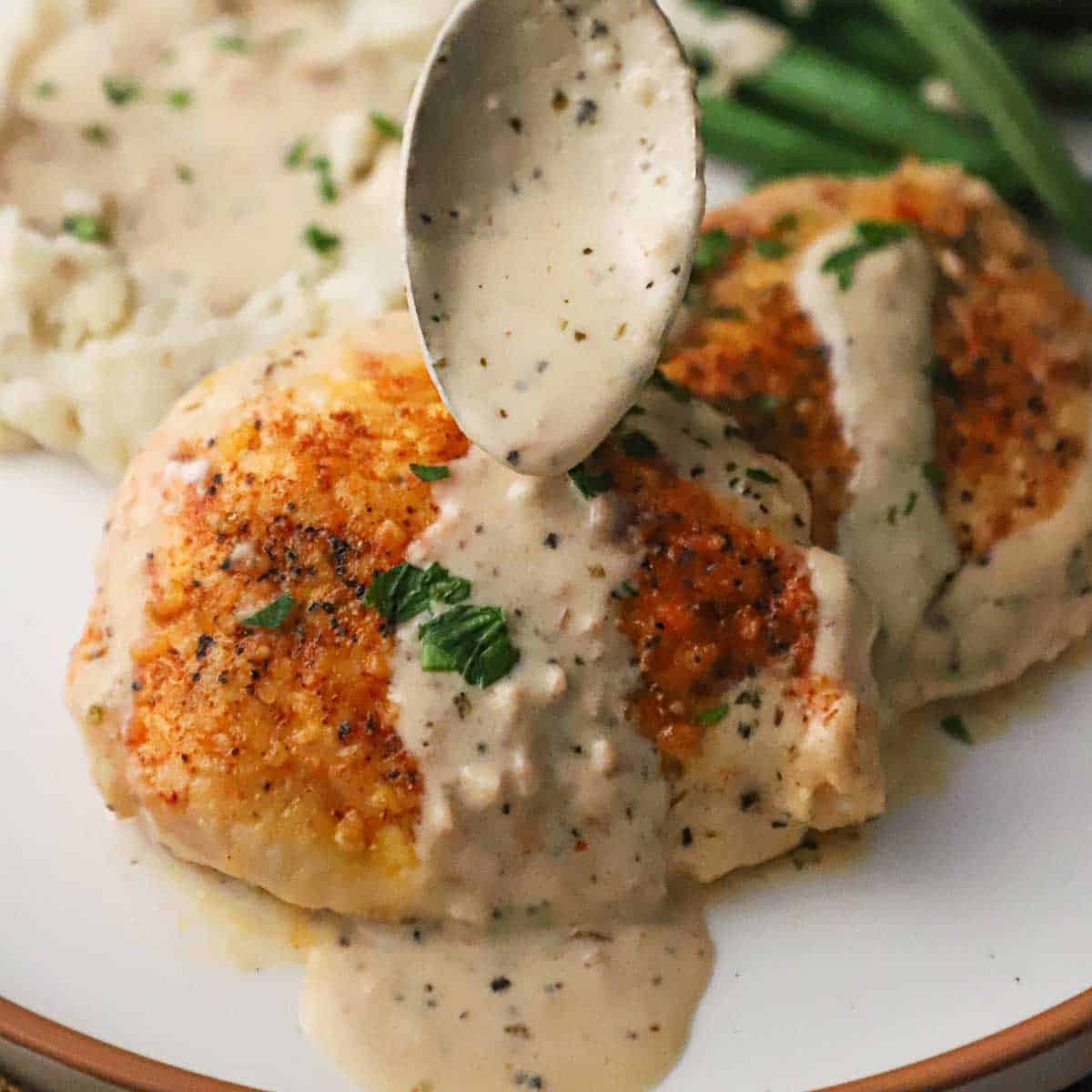 A person using a spoon to drizzle a garlic Parmesan sauce over the top of a seared boneless chicken breast on a white dinner plate.
