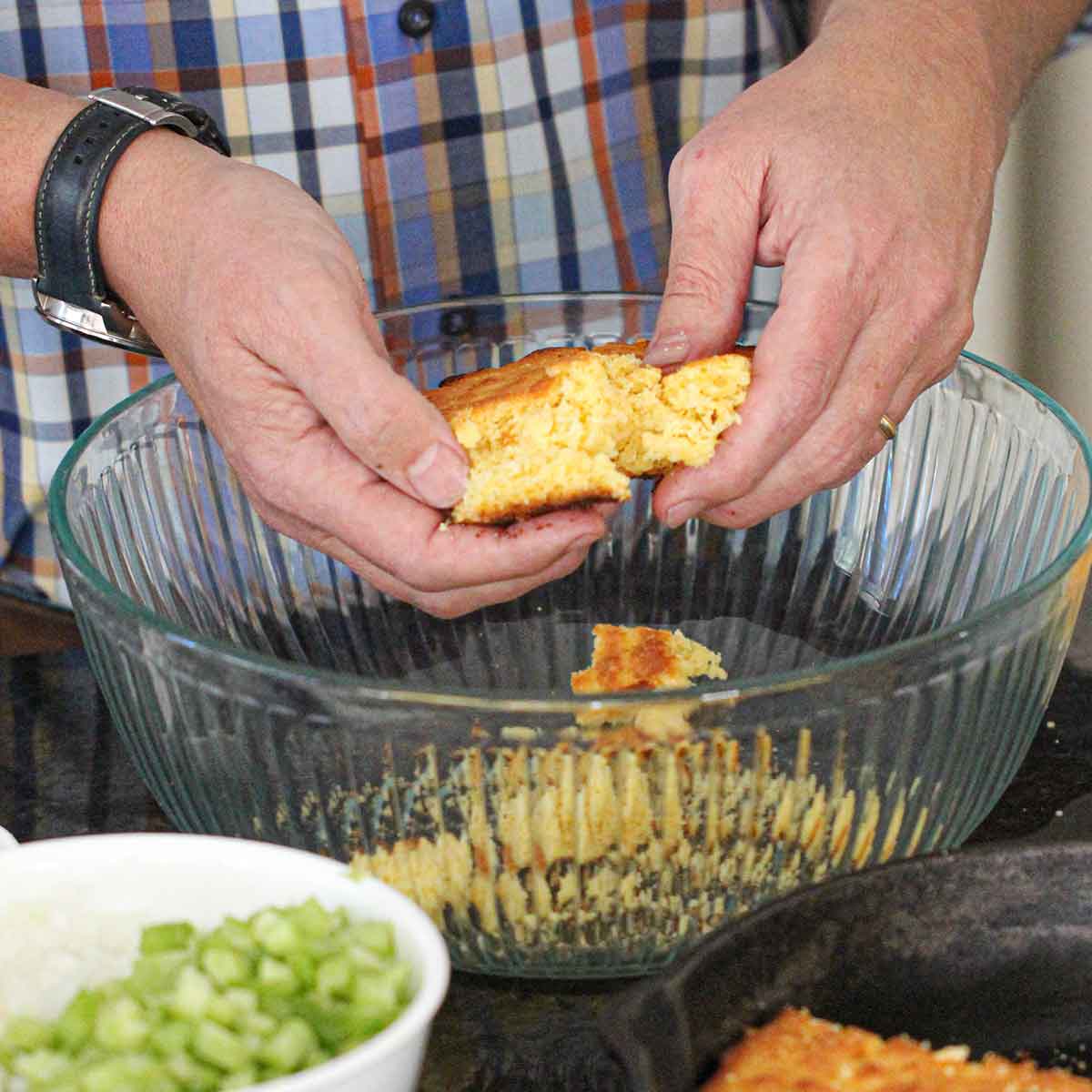 A person using his hands to crumble prepared cornbread in a large glass bowl.