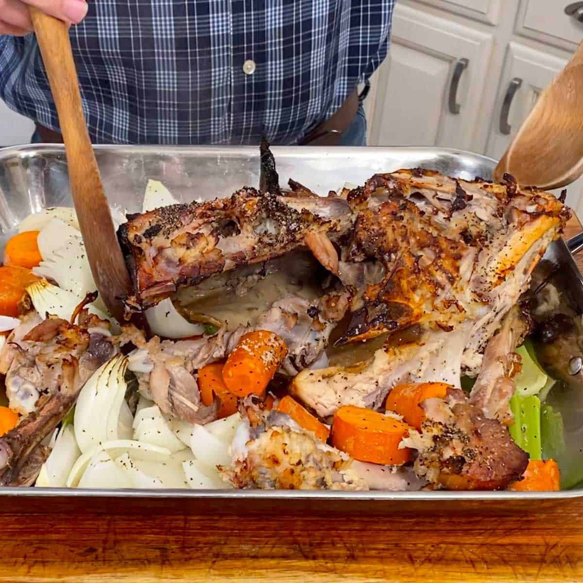 A person using two wooden spoons to toss a turkey carcass and vegetables that have been partially roasted in a hot oven.