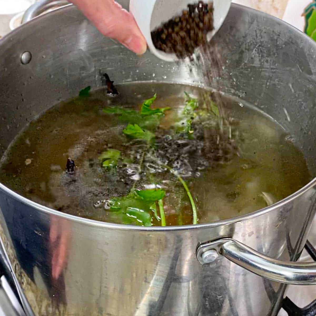 A person transferring black peppercorns from a small white bowl into a large stock pan filled with roasted turkey bones, vegetables, herbs, and simmering water.