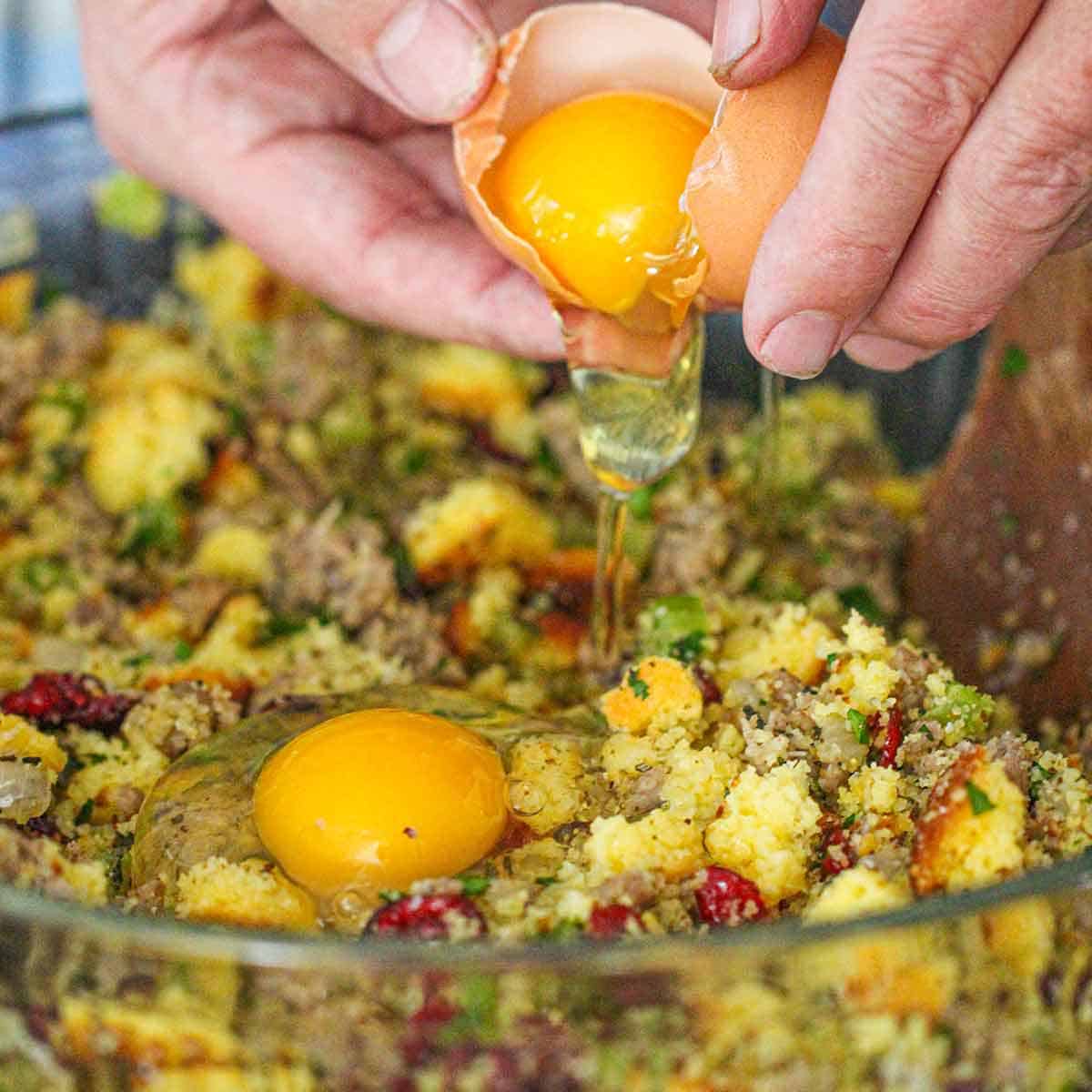 A person cracking a fresh egg into a bowl filled with the ingredients for cornbread dressing with sausage and cranberries.