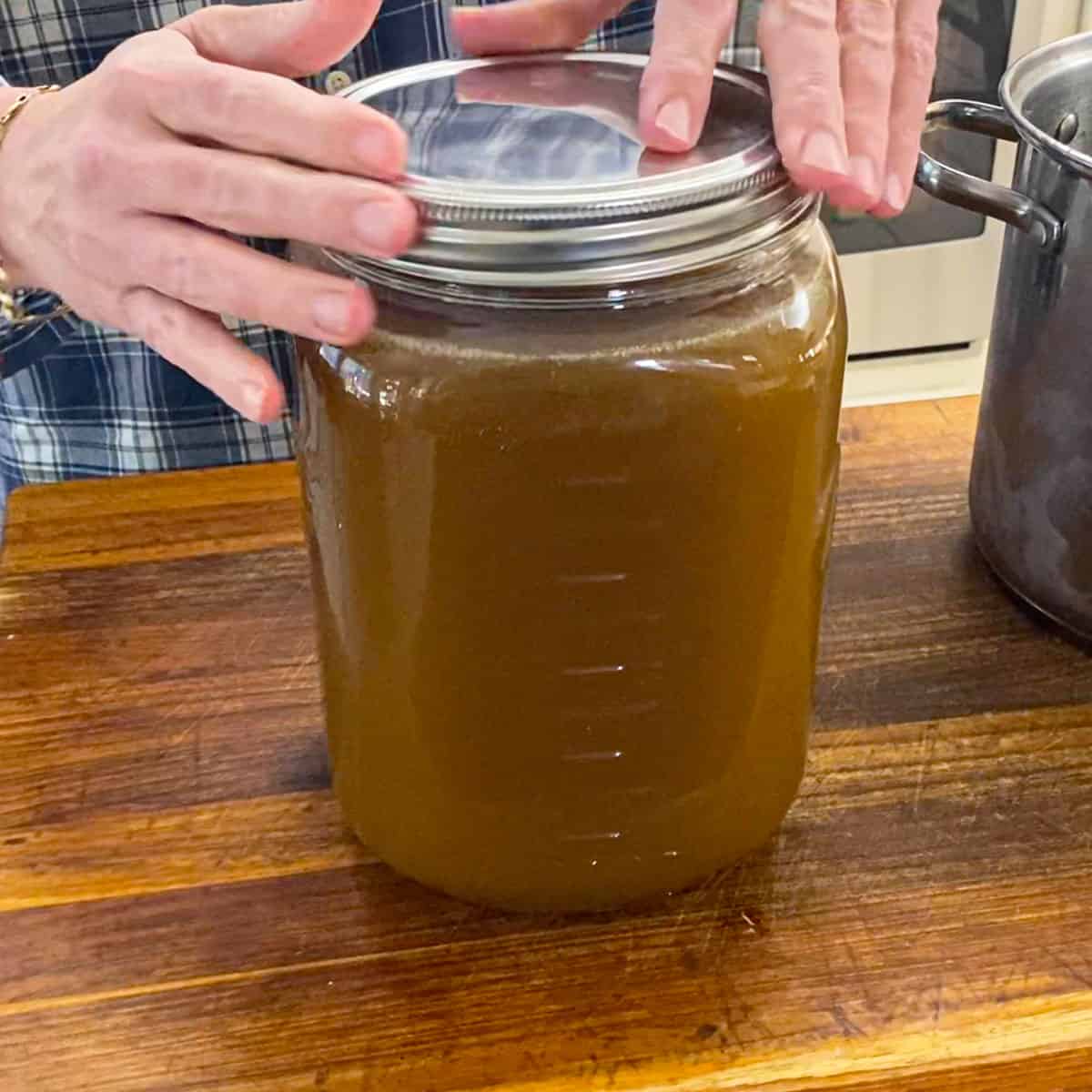 A person twisting on the lid of an oversized jar filled with a gallon of homemade roasted turkey stock on a wooden cutting board.