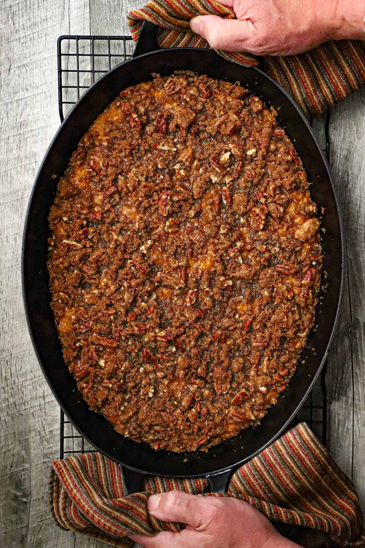 An overhead view of an freshly baked sweet potato casserole with crunchy pecan topping being place on a baking rack by a person using autumnal cloth linens to grip the handles.