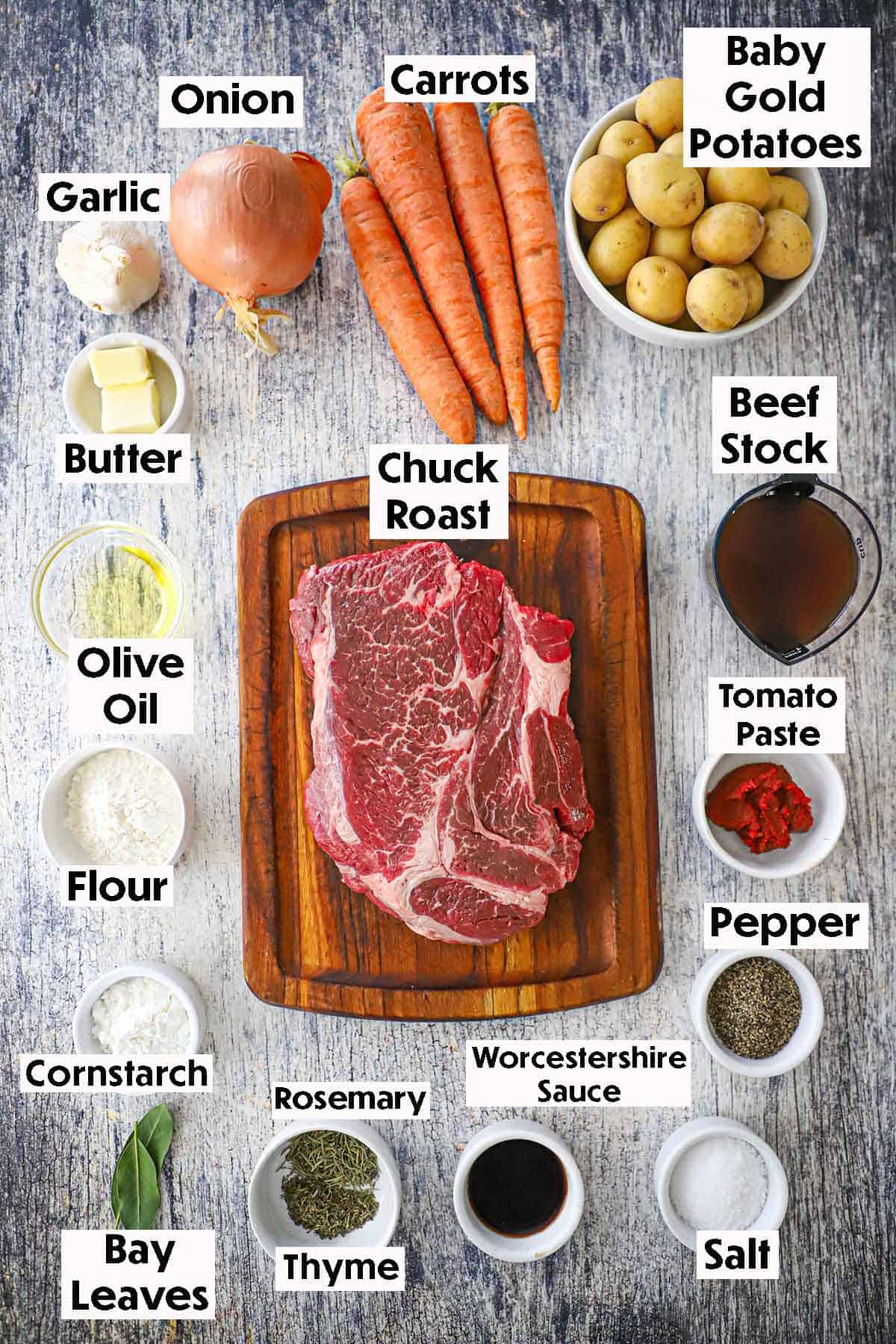 An arrangement of ingredients for the best slow cooker pot roast on a grey wooden background including an uncooked chuck roast, carrots, potatoes, onions, garlic, butter, beef stock, tomato paste, oil, Worcestershire sauce, and seasonings.