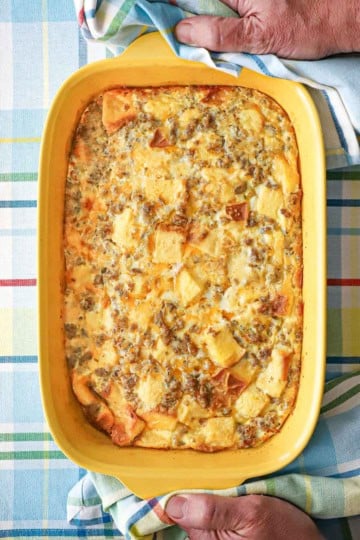 A yellow rectangular baking dish filled with a classic breakfast casserole with the handles of the dish being held by a person holding two linen napkins.