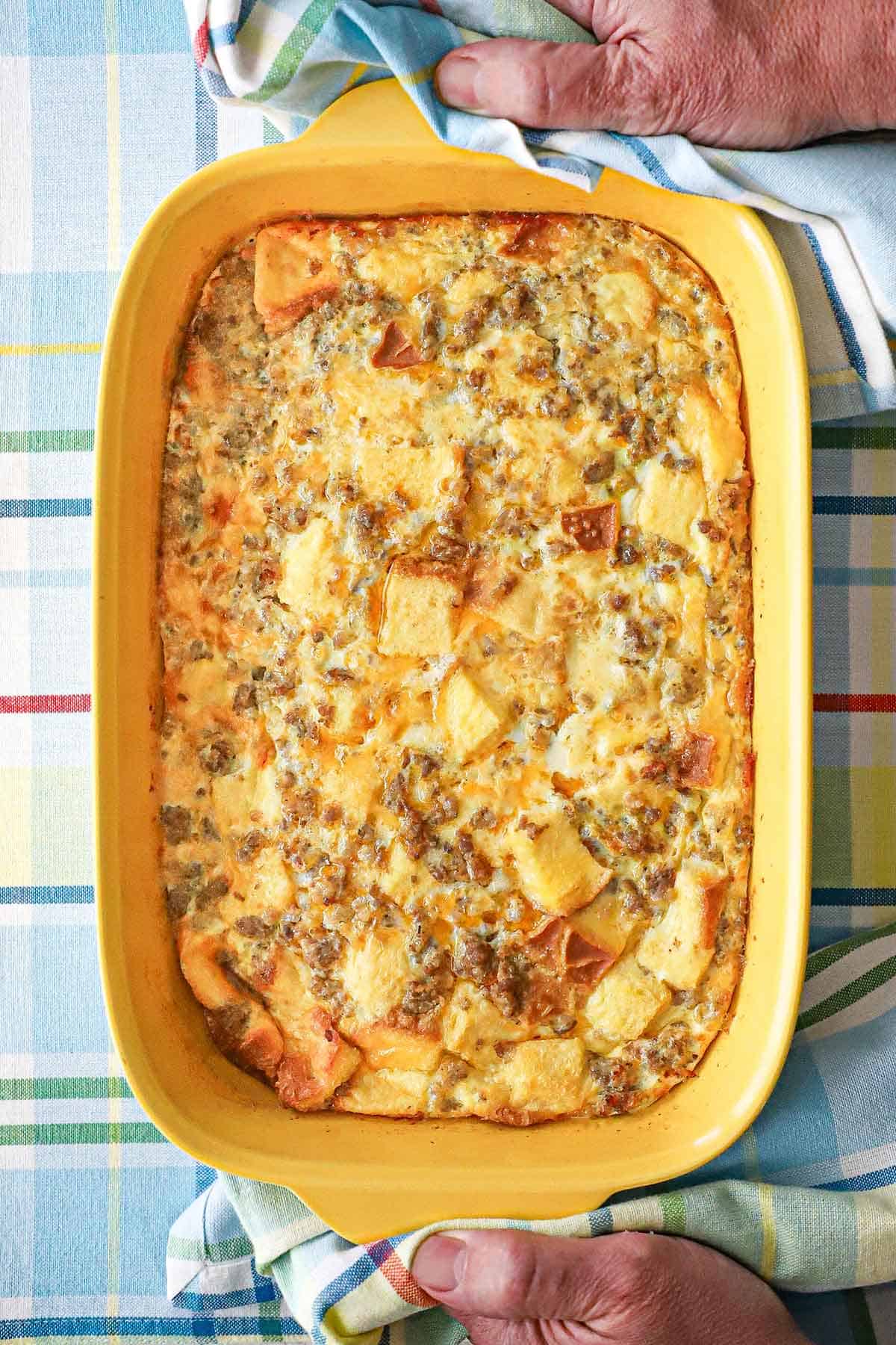 A yellow rectangular baking dish filled with a classic breakfast casserole with the handles of the dish being held by a person holding two linen napkins.
