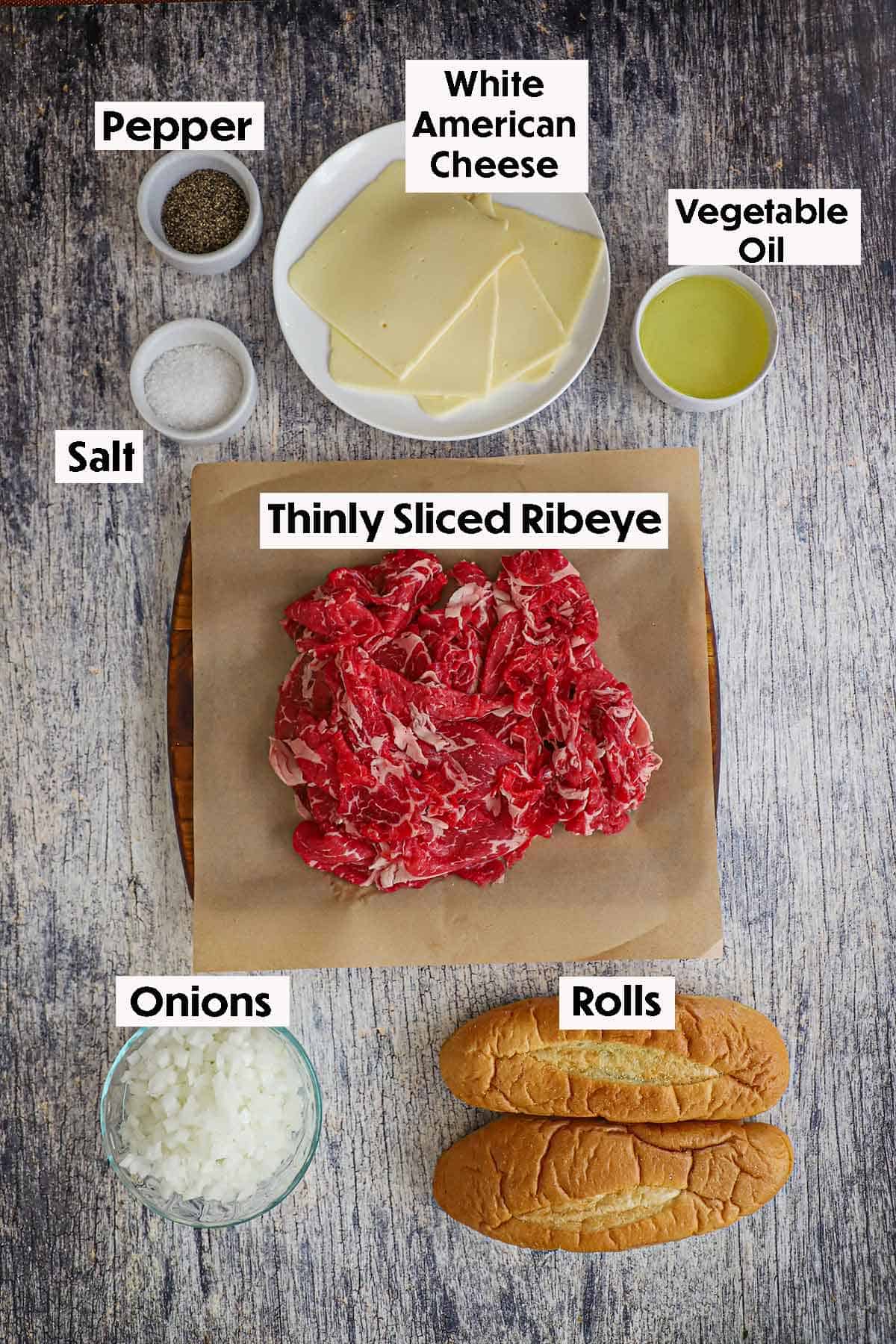 An arrangement of ingredients for homemade Philly cheesesteak on a grey, wooden background inlcuding shave ribey, salt, peper, oil, chopped onions, and rolls. 
