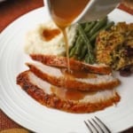 A person pouring turkey gravy from a white gravy boat over a plate filled with three slices of maple glazed whole turkey breast next to cornbread dressing, green beans, and mashed potatoes with gravy.