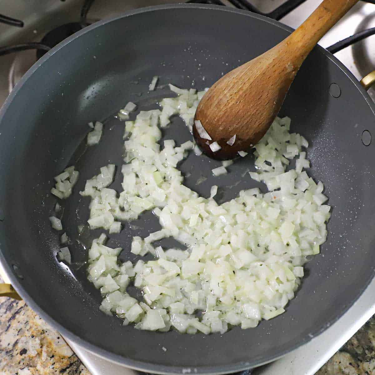 A person using a wooden spoon to stir and sauté chopped onions in melted butter in a large non-stick pan on a gas stove.