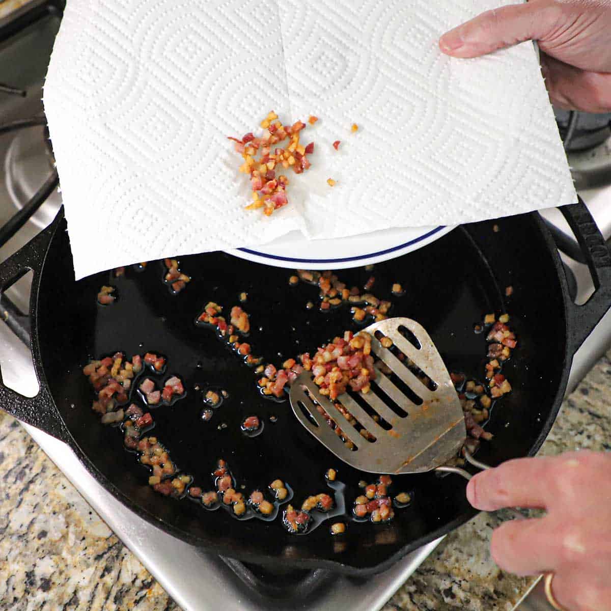 A person using a slotted metal spatula to transfer crispy, cooked pancetta from a black cast-iron skillet onto a paper towel-lined plate.