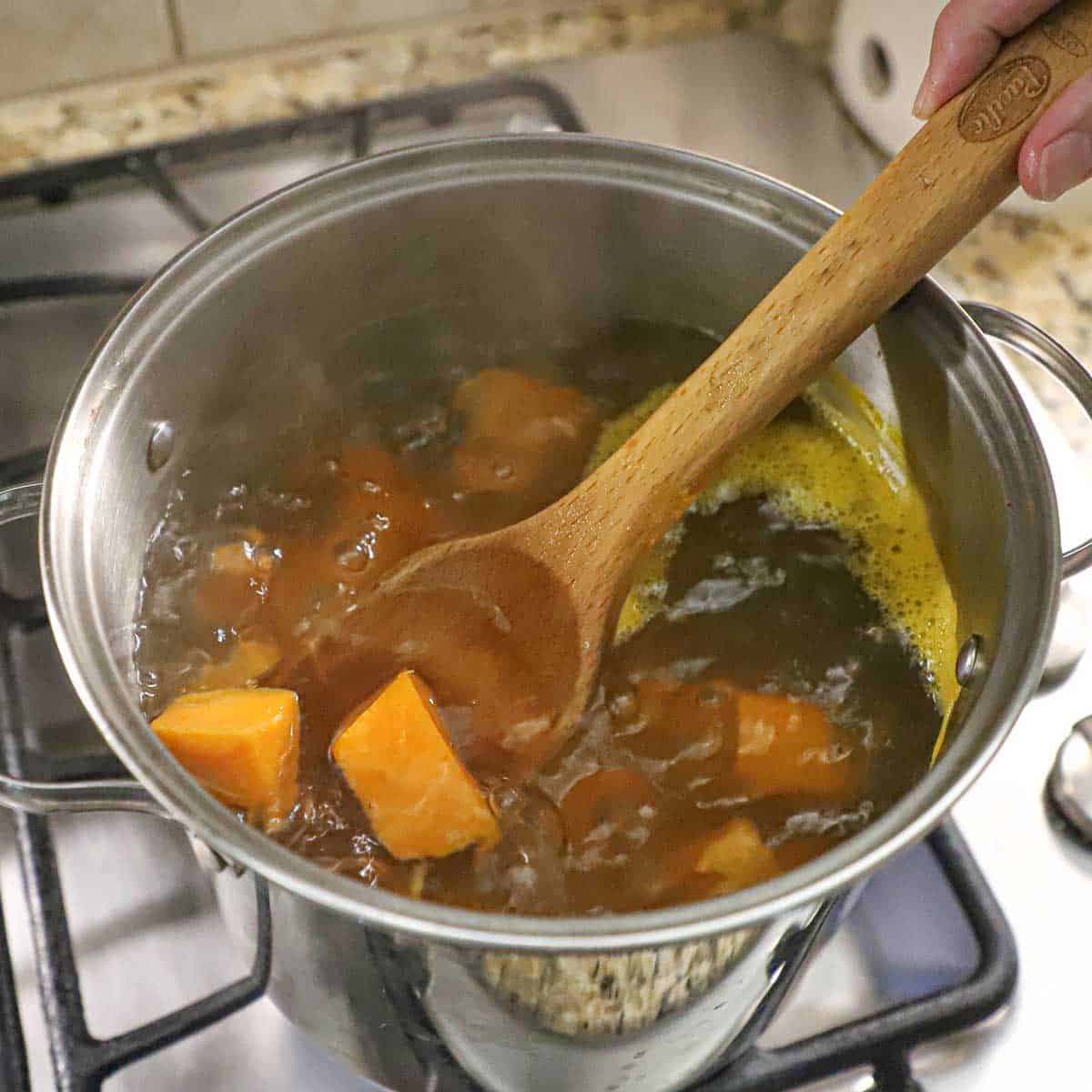 A person stirring cubes of sweet potatoes in a stock pan filled with boiling water on a gas stove.