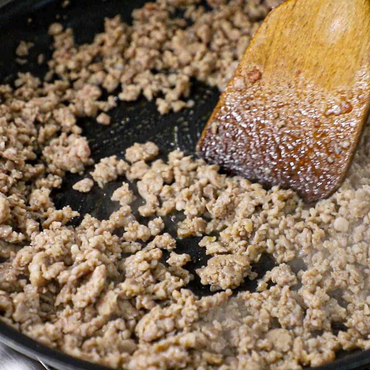 A close-up view of breakfast sausage that is being browned and stirred with a wooden spatula in a black non-stick skillet.