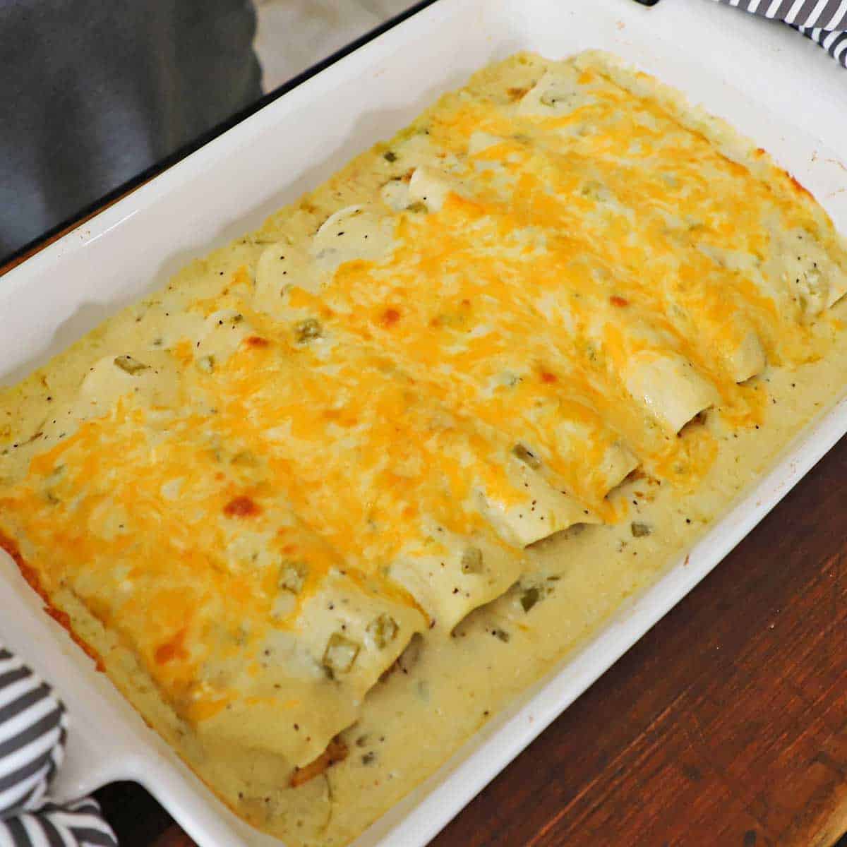 A casserole dish filled with freshly baked chicken enchiladas with a sour cream sauce resting on a cutting board with a person holding the sides with oven mitts.