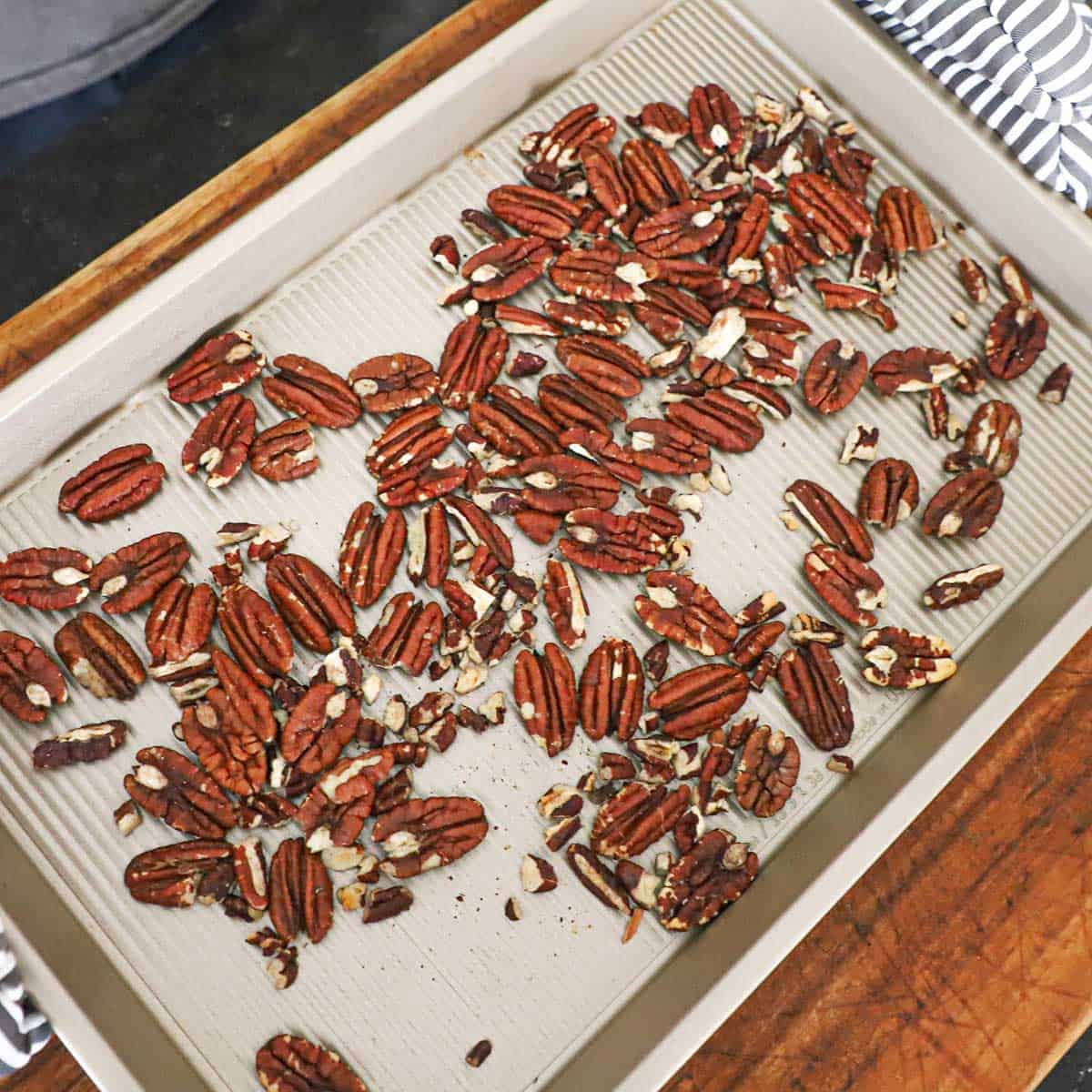 A person holding a small golden baking sheet pan with toasted pecans in the middle of the pan.