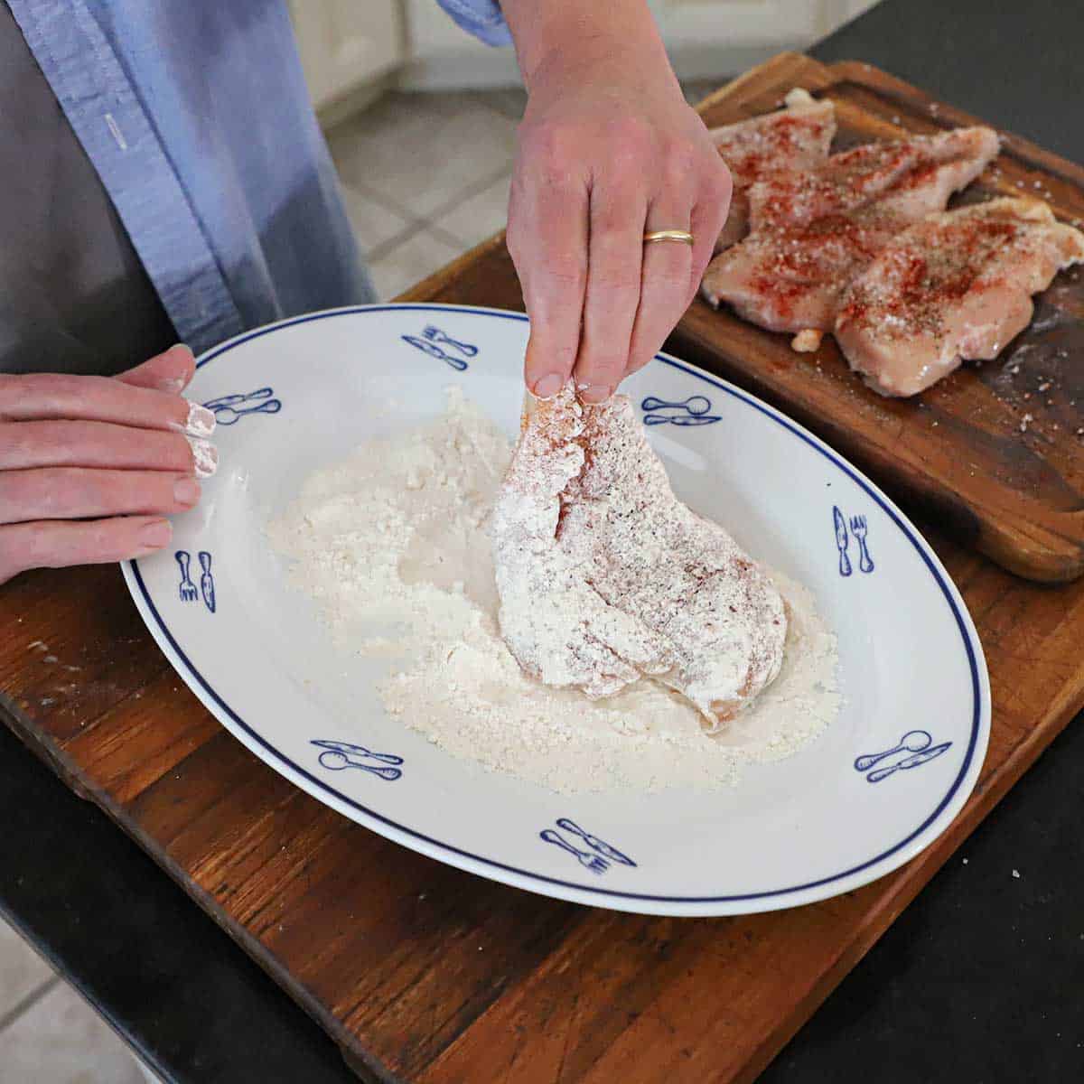 A person dredging a seasoned boneless chicken breast through all-purpose flour on a white platter.