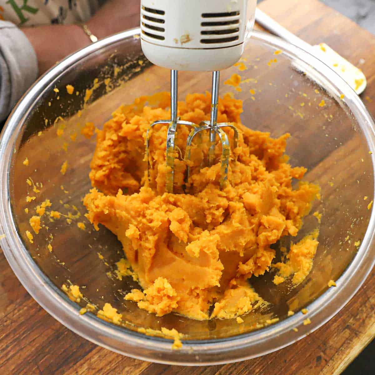 A person using an electric hand mixer to mix boiled sweet potatoes in a glass bowl.