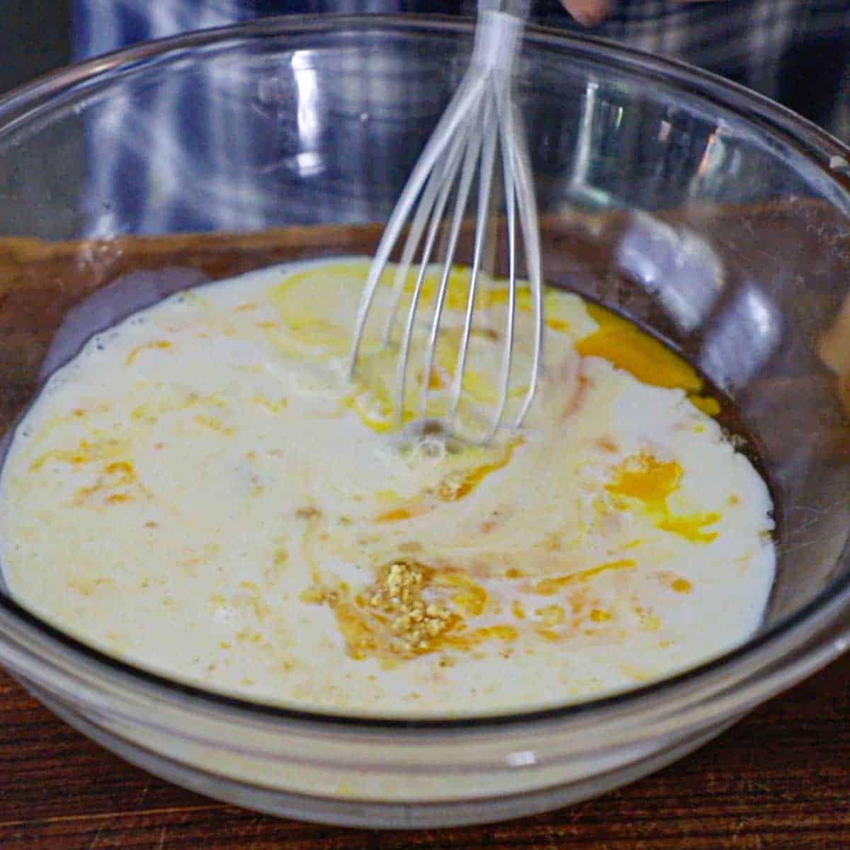 A person using a whisk to mix whole eggs, milk, salt, and dry mustard in a large glass bowl on a wooden cutting board.