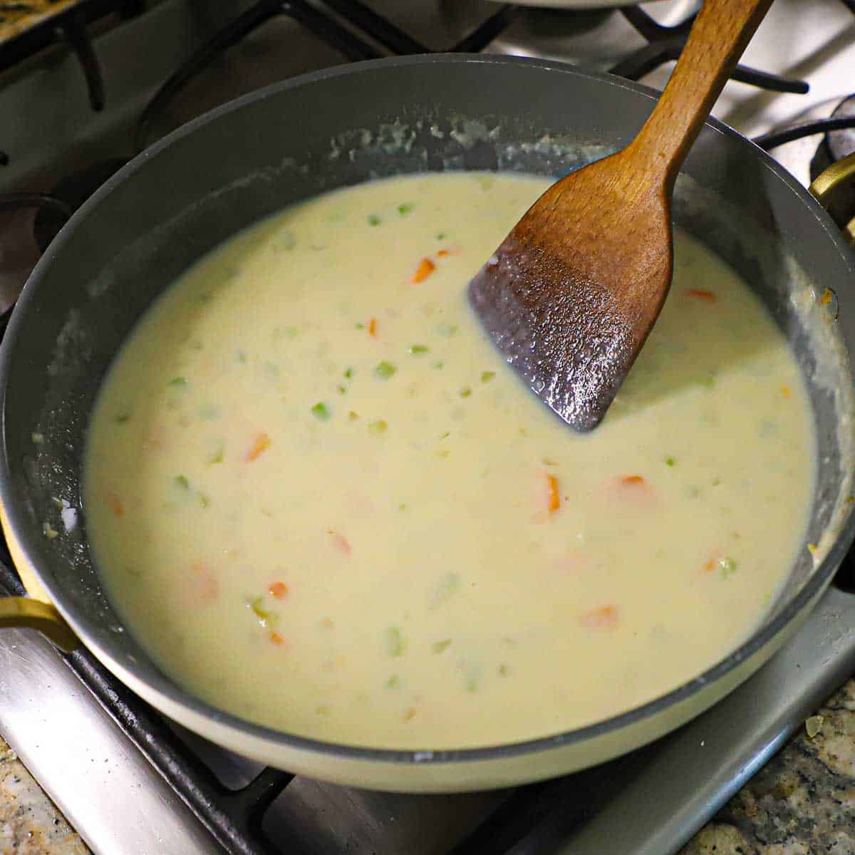 A person using a wooden spatula to stir a creamy sauce of chicken broth, milk, and sautéd vegetables.
