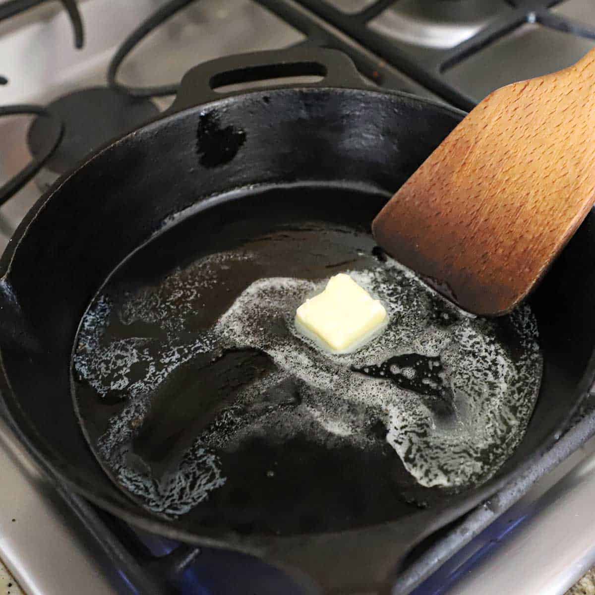 A person using a large wooden spatula to stir a pad of butter that is melting in a cast-iron skillet on a gas stove.