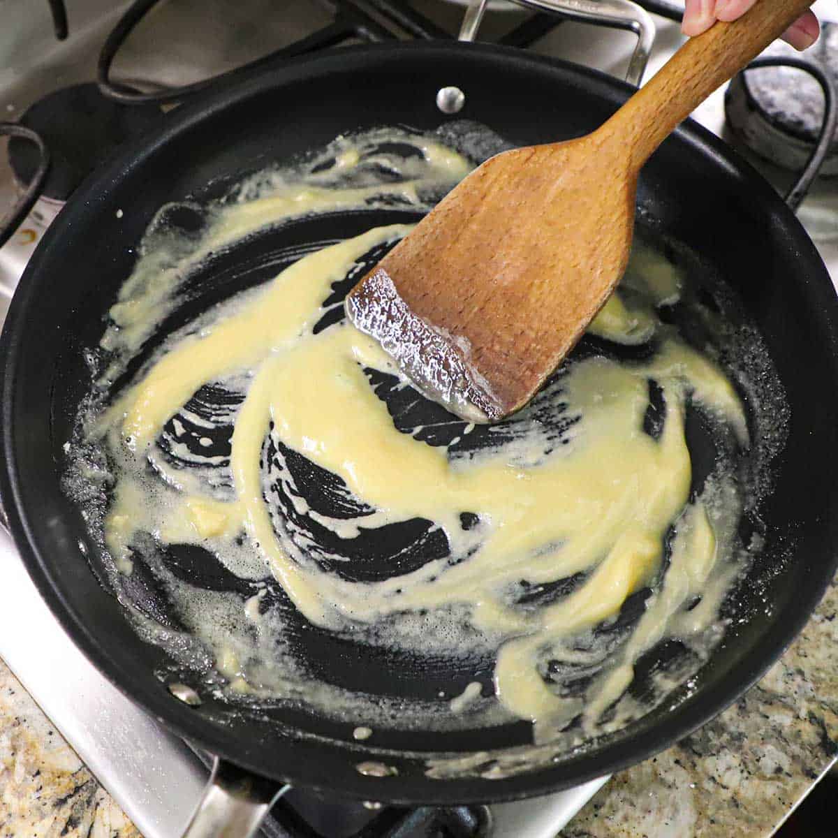 A person using a wooden spatula to stir flour into melted butter to create a roux in a large black non-stick skillet on a gas stove.