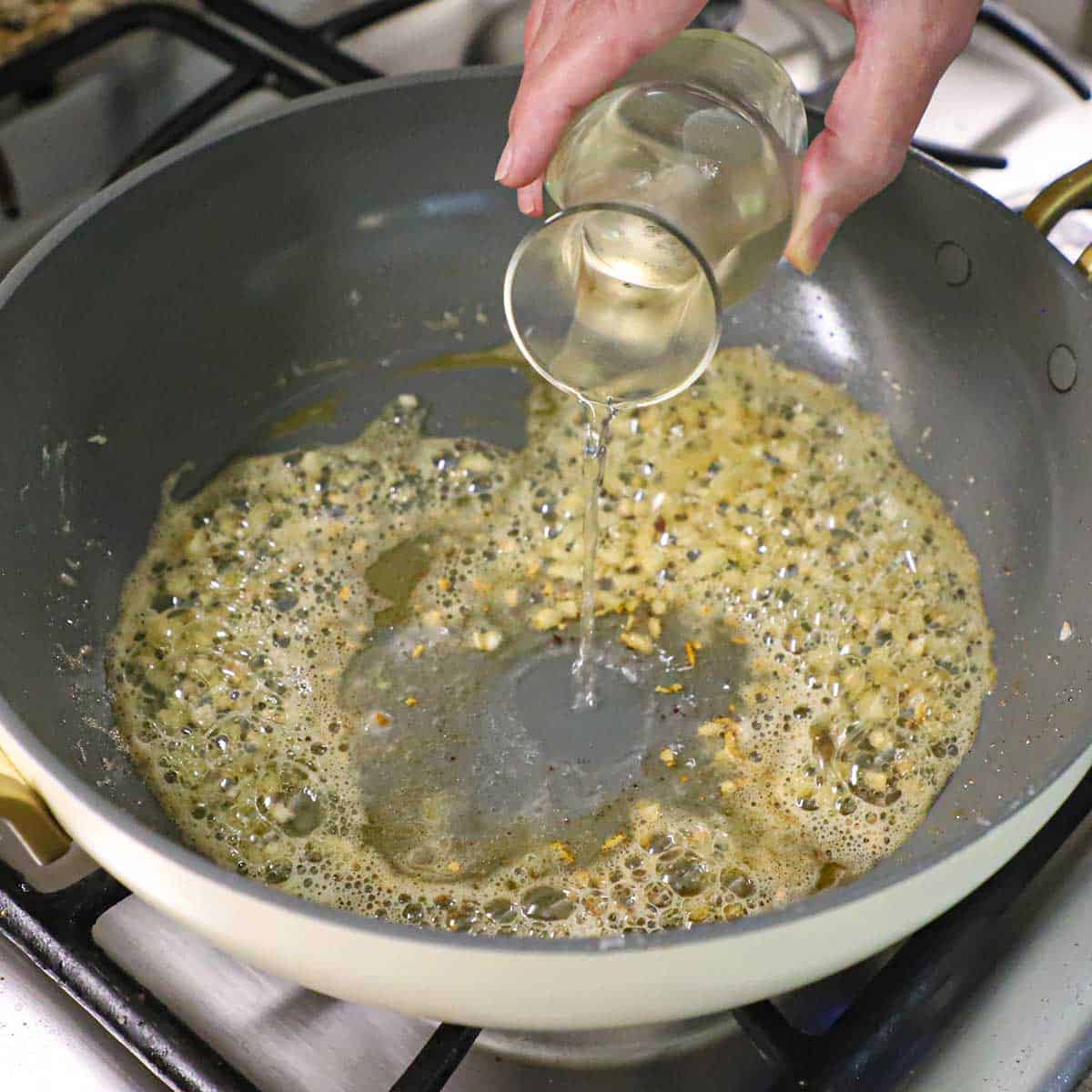 A person pouring white wine from a small carafe into a skillet filled with simmering oil with minced, sautéed garlic in it.