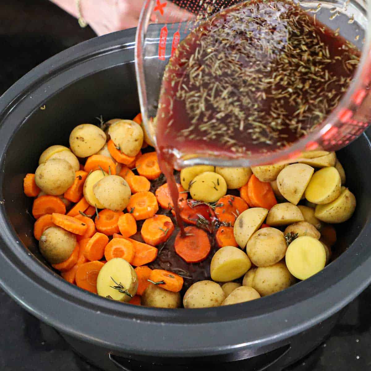 A person pouring a red wine and beef stock with herbs mixture into a slow cooker filled with a seared pot roast and sliced carrots and potatoes.
