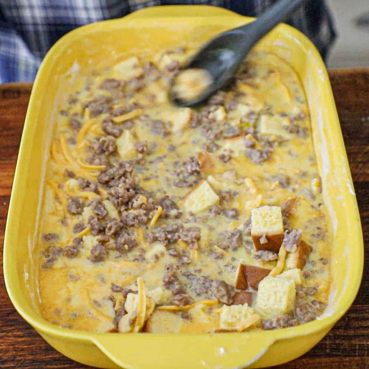 A person using a grey plastic spoon to smooth an uncooked mixture of classic breakfast casserole in a 9 by 13 inch yellow casserole dish.