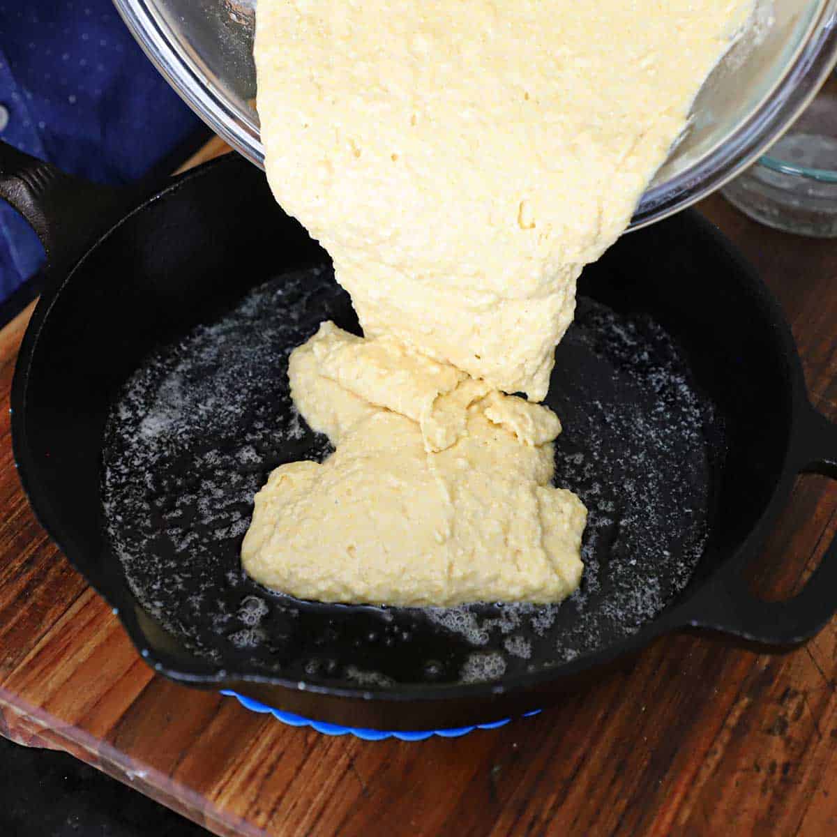 A person transferring cornbread batter from a large glass bowl into a cast-iron skillet that has a layer of melted butter in the bottom of it.