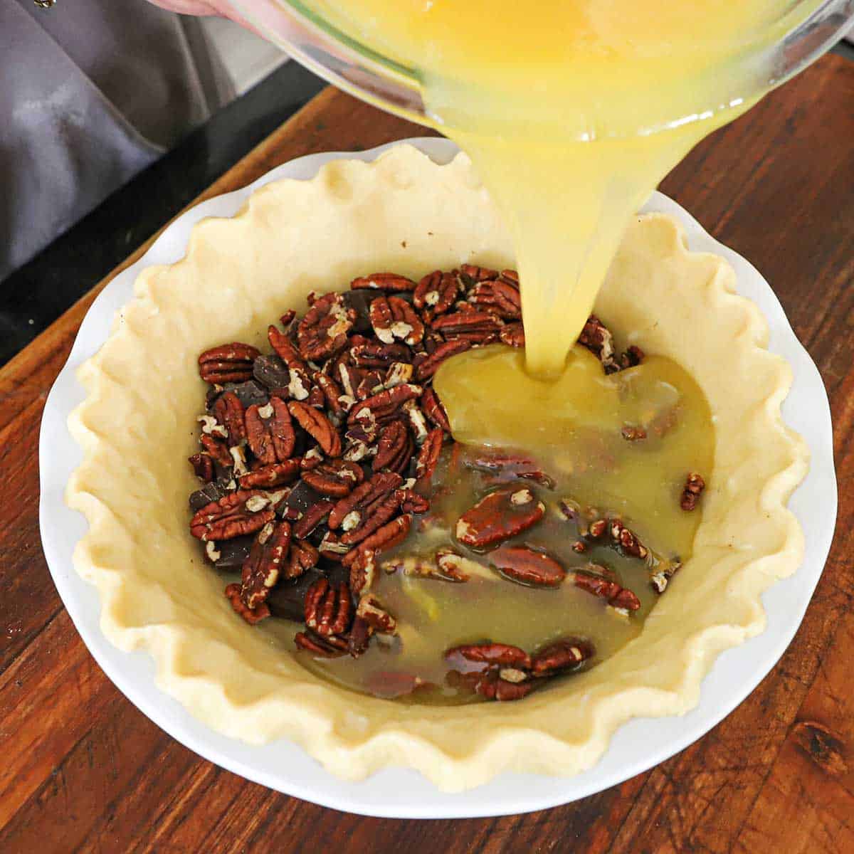 A person pouring pecan pie filling over pecan halves and chunks of chocolate in an the bottom of an uncooked pie dough that has been fitted into a white pie dish.