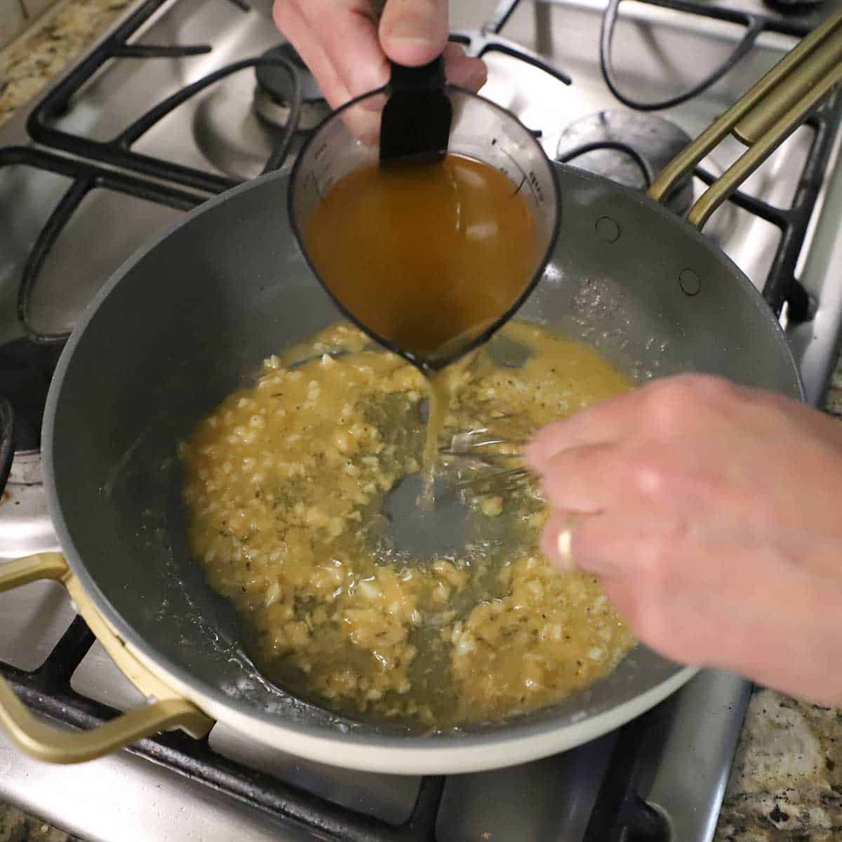 A person pouring chicken broth from a measuring cup into a skillet that has a roux and sautéed garlic in while using is other hand to whisk the mixture.