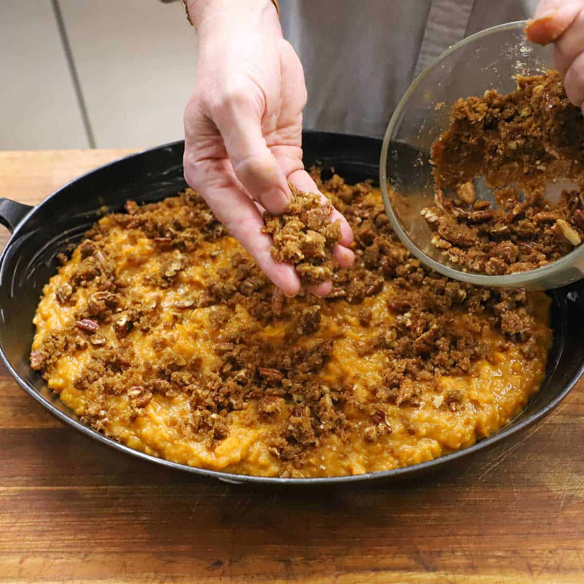 A person sprinkling a brown sugar and chopped pecan mixture all over the surface of an uncooked sweet potato casserole in an oval baking dish.