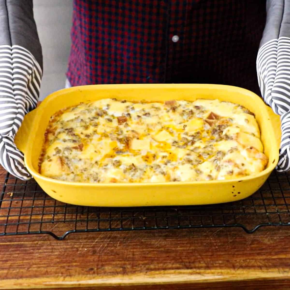 A person using two striped oven mitts to hold a 9 by 13 yellow casserole dish that is filled with classic breakfast casserole that has just come out of the oven.