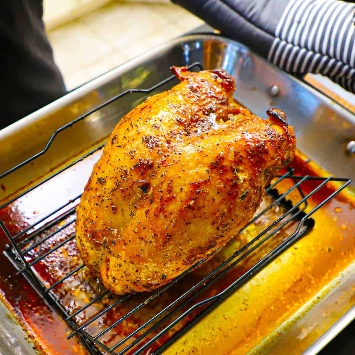 A person wearing oven mitts to hold a roasting pan that is filled with a maple glazed turkey breast on a baking rack fitted inside a stainless steel roasting pan.