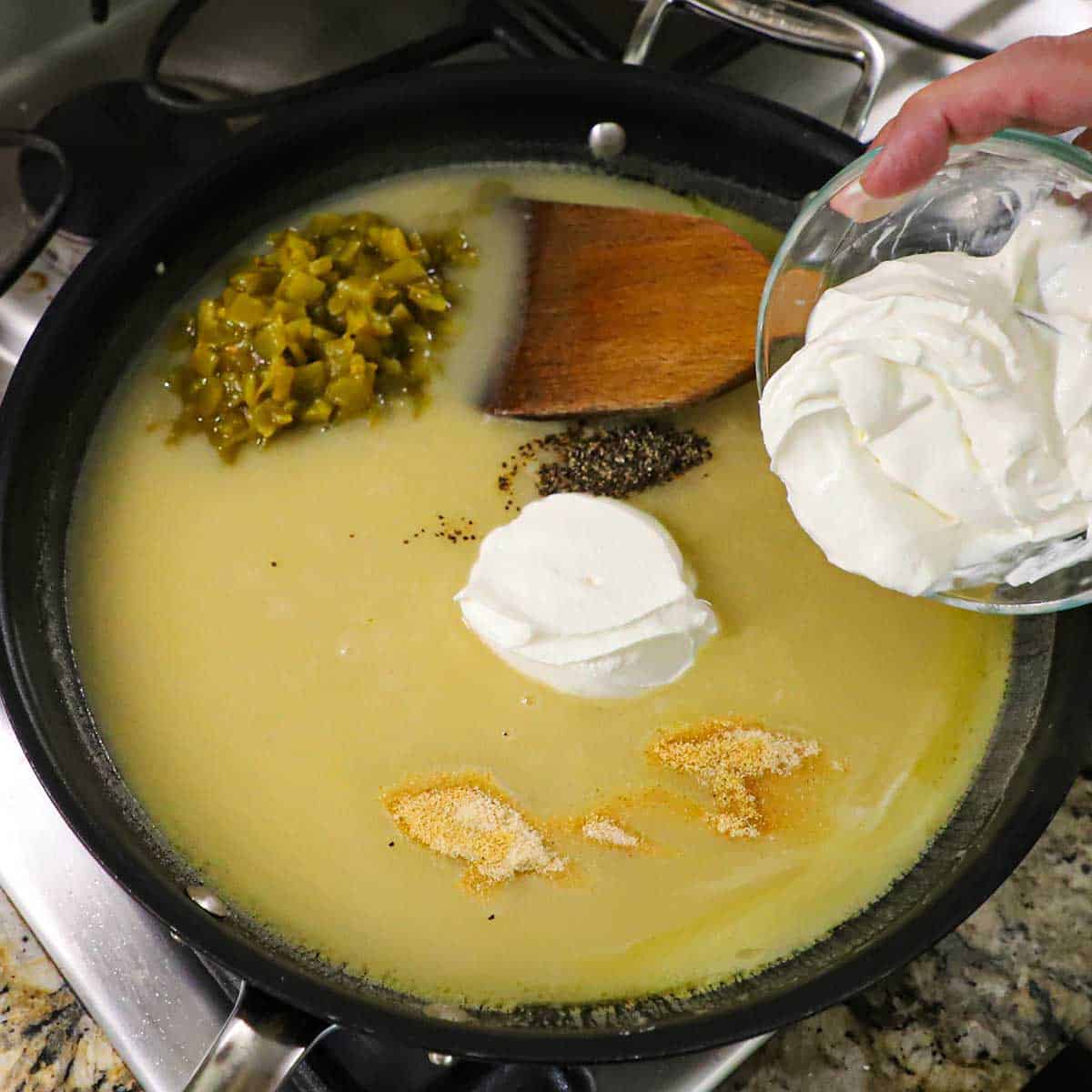 A person transferring sour cream from a glass bowl into a skillet filled with a thicken chicken broth along with green chiles, onion and garlic powder, and ground black pepper.