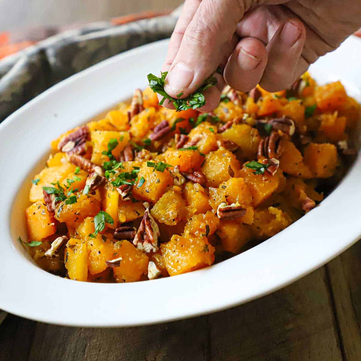 A person using his fingers to garnish a serving bowl filled with maple-glazed butternut squash with chopped parsley and chopped pecans.