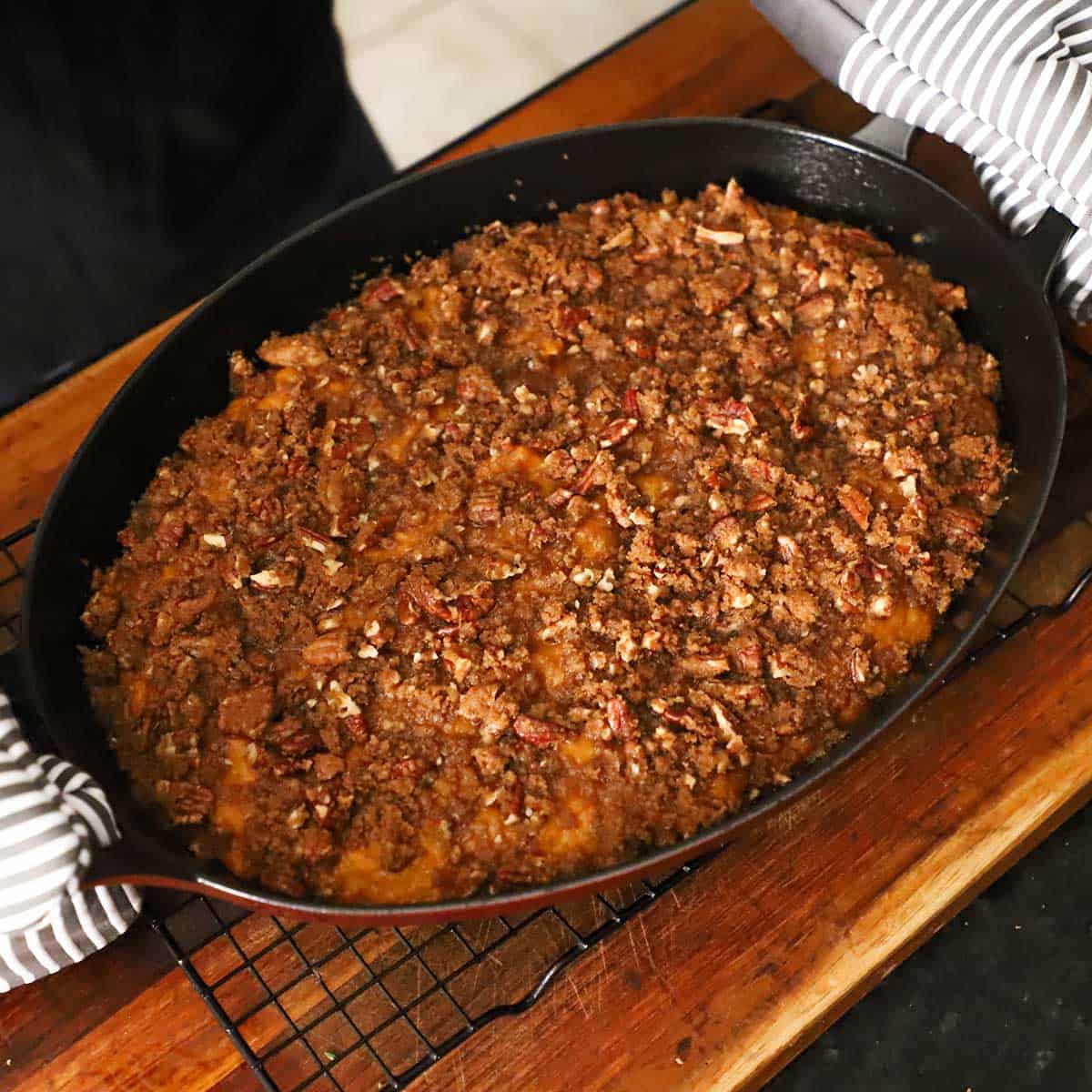 A person using a pair of oven mitts to place an oval baking dish filled with a freshly baked sweet potato casserole with crunch pecan topping on a baking rack on a cutting board.