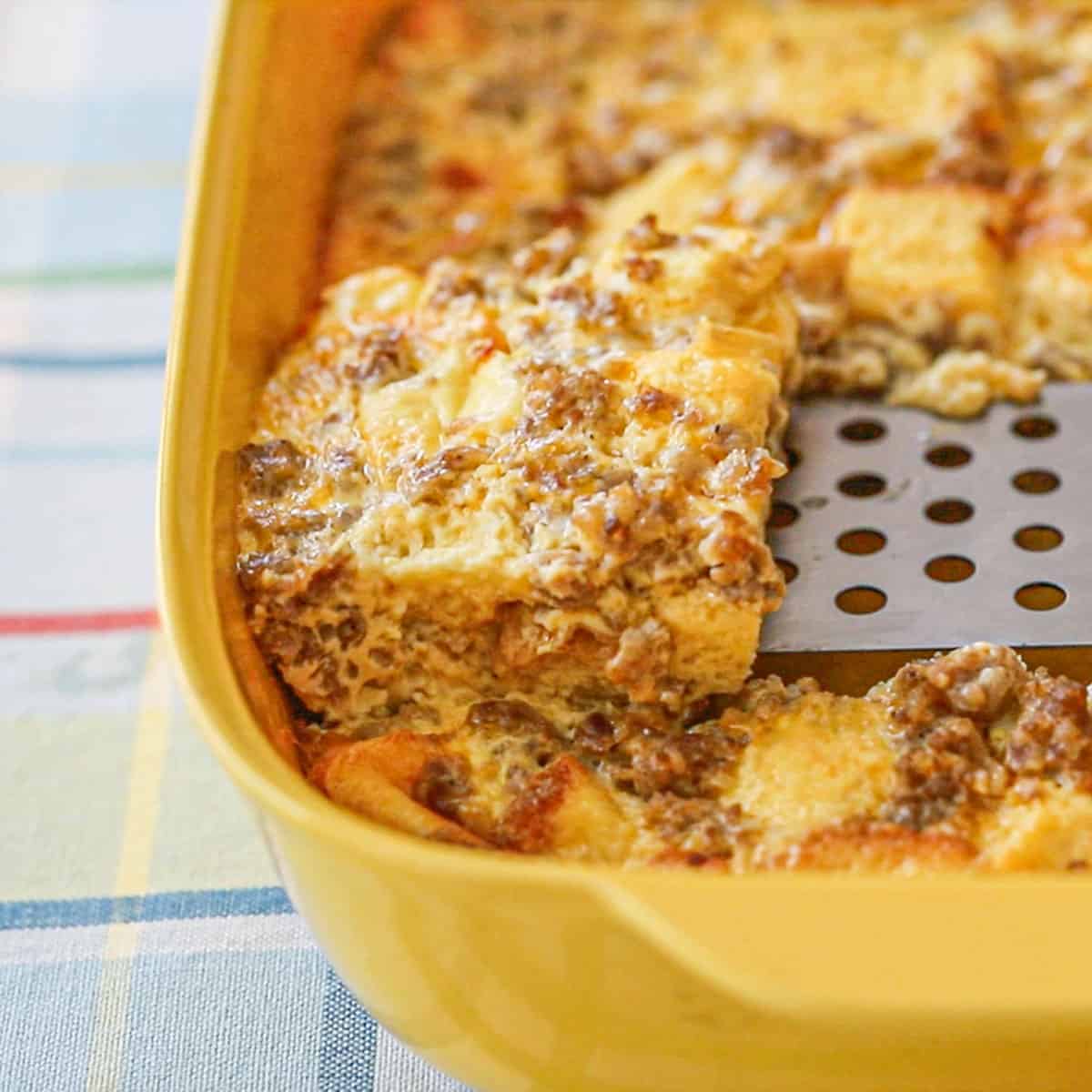 A person using a metal spatula to lift a slice of classic breakfast casserole from a dish that is filled with the casserole.
