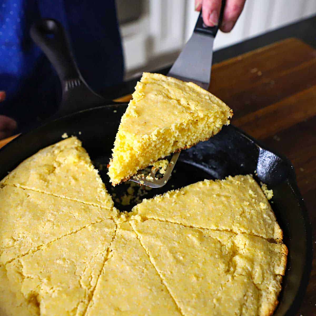 A person using a small spatula to lift a triangular slice of homemade cornbread from a black cast-iron skillet filled with more slices of the cornbread.