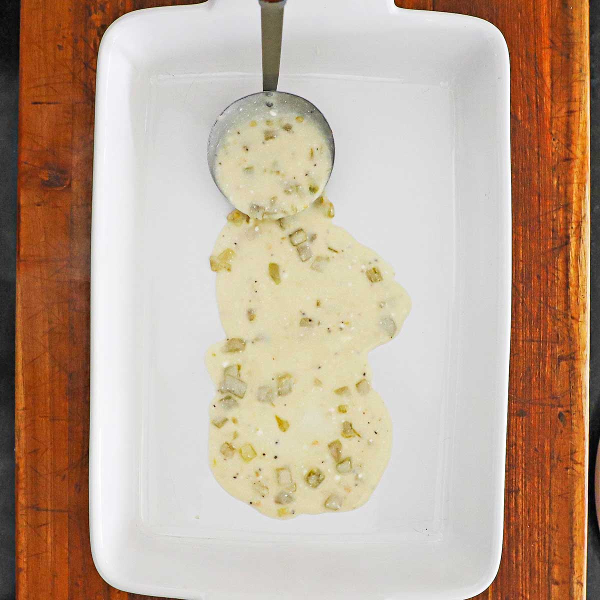 A person using a small ladle to spread a thin layer of a sour cream sauce over the bottom of a white casserole dish.