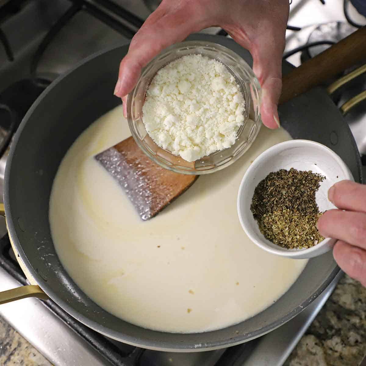 A person holding a small bowl of grated parmesan cheese in one hand and a small bowl of dried herbs in his other hand, both over a skillet filled with a silky cream sauce.