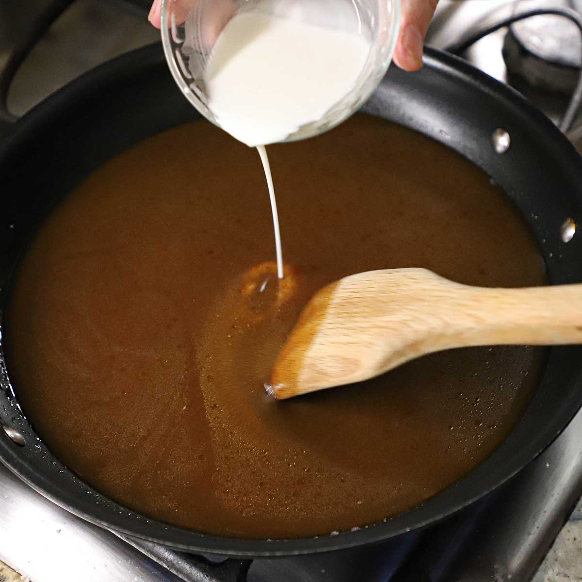A person pouring a cornstarch slurry into a brown gravy that is being stirred and simmered in a large black non-stick skillet.